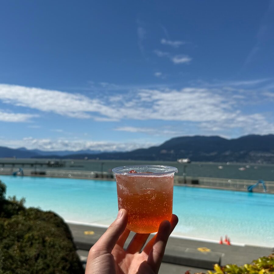 A cup of orange Sprizzini held up in front of Kitsilano Pool and the North Shore mountains